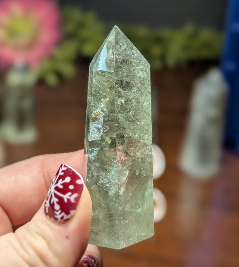 May include: A pale green crystal point with a faceted, pointed top. The crystal is held in a hand with a red and white snowflake design on the fingernail. The crystal has internal fractures and inclusions.