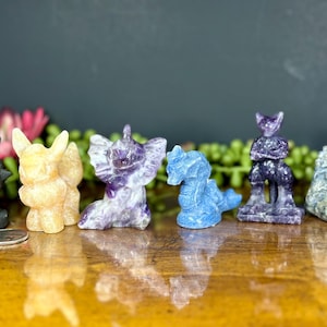 May include: A collection of seven small, hand-carved crystal animal figurines. The figurines are in various colors, including black, orange, purple, blue, and gray. The figurines are arranged in a row on a wooden surface.
