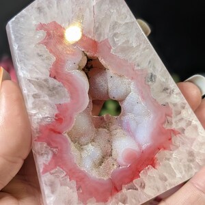May include: A polished, crystalline agate geode with a pink and white interior. The geode has a natural, irregular shape with a hollow center. The outer layer is a translucent, light gray color. The geode is held in a hand.