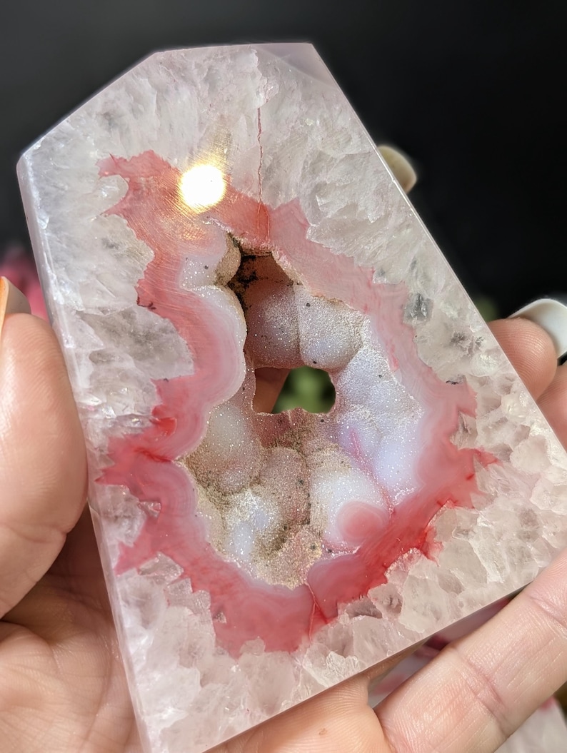 May include: A polished, crystalline agate geode with a pink and white interior. The geode has a natural, irregular shape with a hollow center. The outer layer is a translucent, light gray color. The geode is held in a hand.