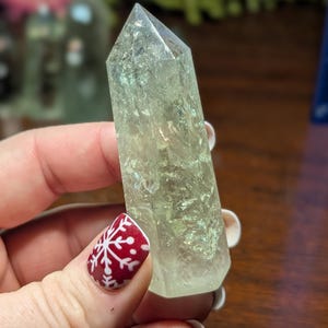 May include: A clear, pale green crystal point with a faceted tip. The crystal is held in a hand with a red and white snowflake nail design. Other crystals are visible in the background.
