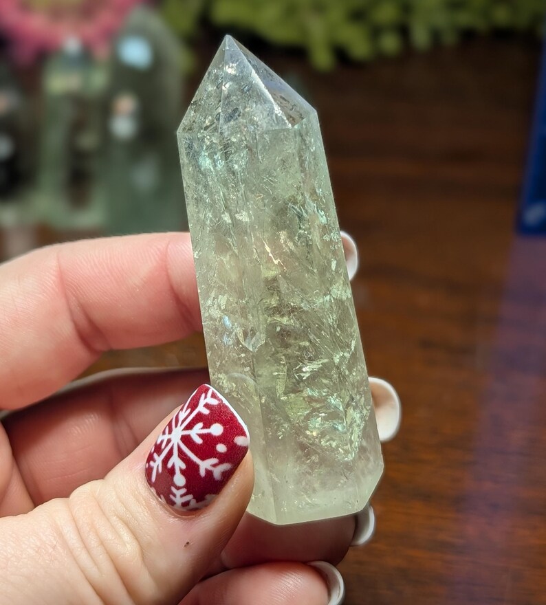 May include: A clear, pale green crystal point with a faceted tip. The crystal is held in a hand with a red and white snowflake nail design. Other crystals are visible in the background.