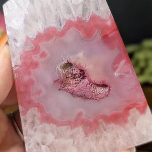 May include: A polished, rectangular crystal with a pink and white agate pattern. The center features a geode-like cavity filled with a cluster of small, sparkling crystals. The crystal's edges are a translucent white, and the interior displays a vibrant pink hue.