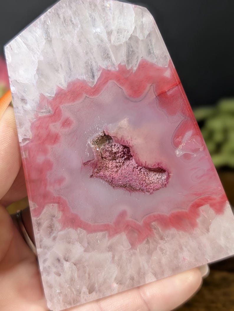 May include: A polished, rectangular crystal with a pink and white agate pattern. The center features a geode-like cavity filled with a cluster of small, sparkling crystals. The crystal's edges are a translucent white, and the interior displays a vibrant pink hue.