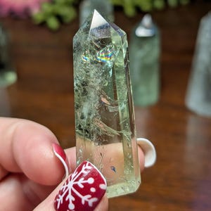 May include: A light green crystal point with internal fractures and rainbow iridescence. The crystal is held in a hand with red nail polish and a white snowflake design. Other crystals are in the background.
