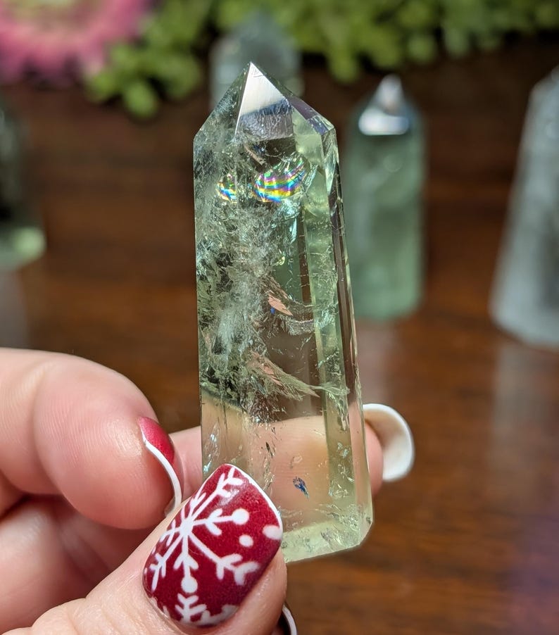 May include: A light green crystal point with internal fractures and rainbow iridescence. The crystal is held in a hand with red nail polish and a white snowflake design. Other crystals are in the background.
