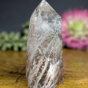 May include: A clear quartz crystal point with internal inclusions and a polished, faceted tip. The crystal has a natural, earthy appearance with streaks of brown and white. The crystal is standing upright on a wooden surface.