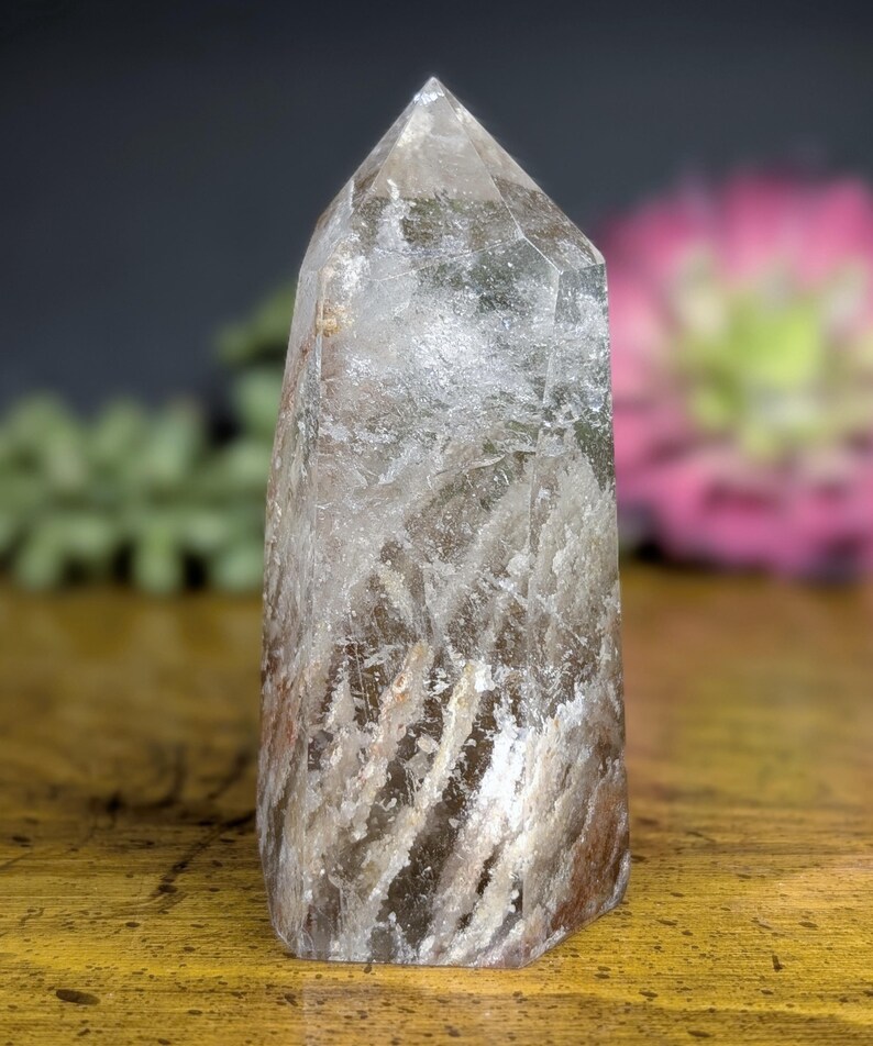 May include: A clear quartz crystal point with internal inclusions and a polished, faceted tip. The crystal has a natural, earthy appearance with streaks of brown and white. The crystal is standing upright on a wooden surface.