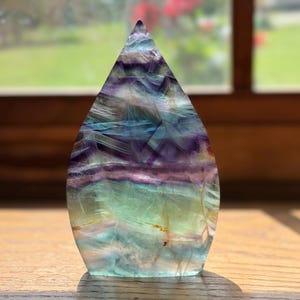 May include: A teardrop-shaped fluorite crystal carving featuring horizontal bands of translucent purple, blue, and green hues. The polished stone displays natural internal fractures and color zoning throughout its smooth, pointed structure.