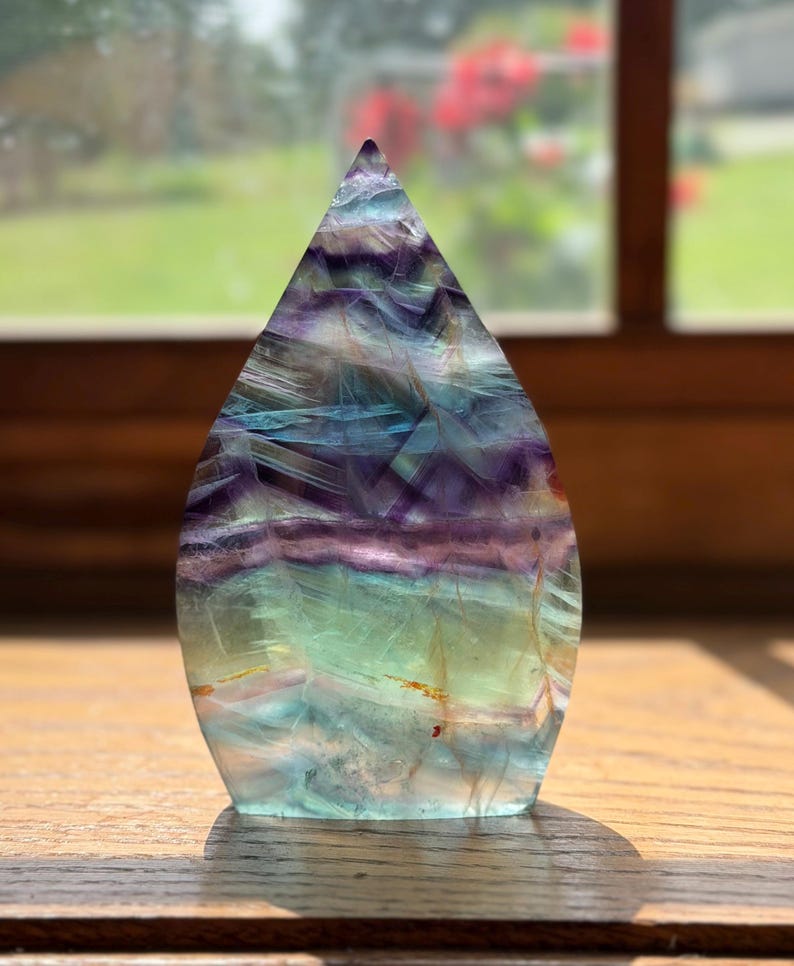 May include: A teardrop-shaped fluorite crystal carving featuring horizontal bands of translucent purple, blue, and green hues. The polished stone displays natural internal fractures and color zoning throughout its smooth, pointed structure.