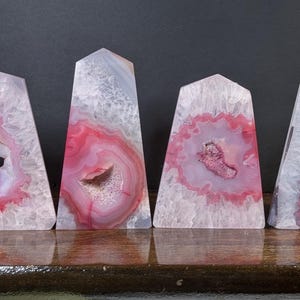 May include: Four agate obelisks with pink and white coloration. Each has a unique pattern of concentric rings and a central cavity. The obelisks are approximately 4 inches tall and stand on a wooden surface.