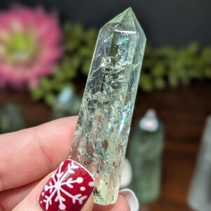 May include: A pale green crystal point with internal fractures and iridescent flashes. The crystal is held in a hand with a red nail with a white snowflake design. Other crystals are visible in the background.