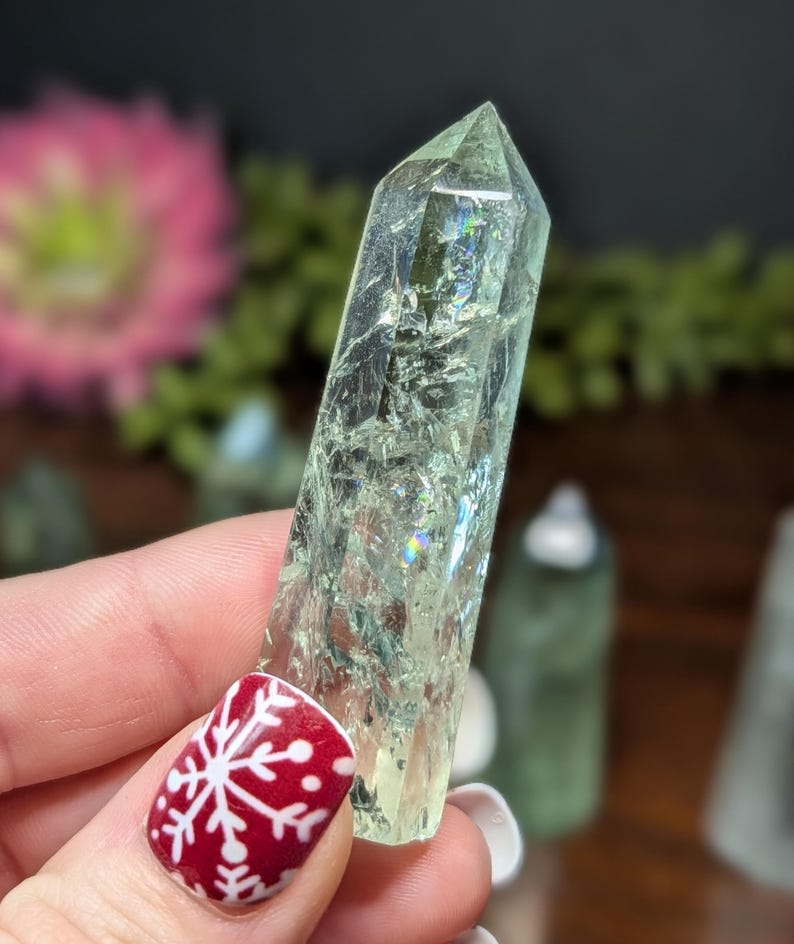 May include: A pale green crystal point with internal fractures and iridescent flashes. The crystal is held in a hand with a red nail with a white snowflake design. Other crystals are visible in the background.