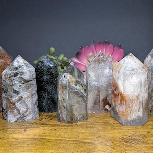 May include: A collection of ten crystal towers in various colors and patterns. The crystals are arranged on a wooden surface with a dark background. The crystals vary in height and have pointed tops. Some crystals have brown, gray, and clear tones.