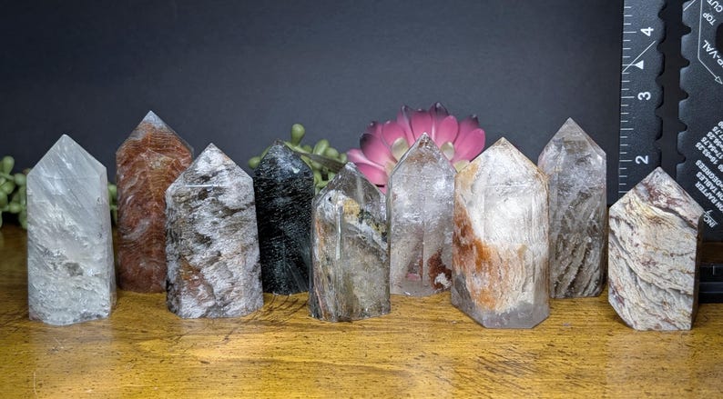 May include: A collection of ten crystal towers in various colors and patterns. The crystals are arranged on a wooden surface with a dark background. The crystals vary in height and have pointed tops. Some crystals have brown, gray, and clear tones.