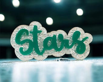 May include: A decorative pin with the word "Stars" in green cursive lettering. The letters are set against a cloud-shaped background with a silver, textured pattern. The pin is on a light blue surface.