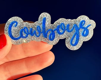 May include: A glittery, silver-colored Cowboys hair clip with blue cursive lettering. The clip is held against a dark blue background. The word "Cowboys" is written in a stylish, flowing font.