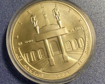 May include: Gold-colored commemorative coin with the inscription "Liberty" and "In God We Trust" on the obverse. The reverse features a stylized depiction of the Los Angeles Coliseum with the inscription "Los Angeles 1984 XXIII Olympiad".