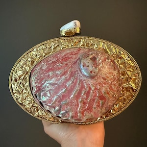 May include: An oval-shaped clutch with a gold-colored frame and a pearlescent, pink and white shell center. The frame has a textured design. A small, white stone sits atop the clutch, attached to a gold-colored clasp.
