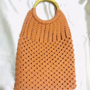 May include: A crocheted tote bag with a brown wooden handle. The bag is made with a light brown yarn and has a woven pattern.