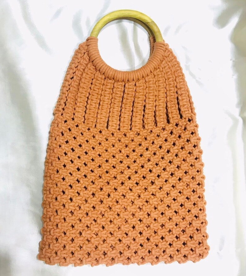 May include: A crocheted tote bag with a brown wooden handle. The bag is made with a light brown yarn and has a woven pattern.