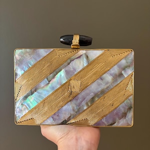 May include: A rectangular clutch bag featuring alternating diagonal panels of iridescent mother-of-pearl and gold-toned wire detailing. The top closure consists of a dark, smooth, cylindrical stone accent mounted on a gold-toned metal frame.