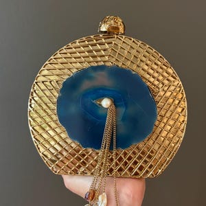 Può includere: Una pochette circolare in metallo dorato con un motivo a griglia testurizzato e una fetta di agata blu al centro. Una piccola perla bianca è incastonata al centro della pietra, con diverse delicate catenine dorate e piccoli ciondoli colorati.