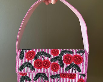 May include: A pink beaded handbag with a cherry design. The bag features a pink and white striped pattern, with red cherries and green leaves. The handle is a solid pink color.