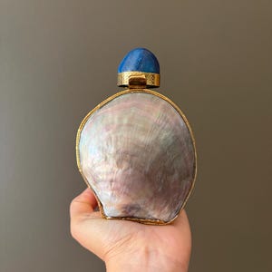 Peut inclure: Une bouteille décorative composée d'un grand corps en nacre ovale et irisé, enchâssé dans un cadre en métal doré. Le sommet de la bouteille est surmonté d'un bouchon en pierre bleu profond, lisse et arrondi, serti dans du métal doré.
