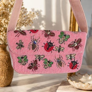 May include: A pink beaded handbag featuring a variety of embroidered insect motifs, including ladybugs, beetles, and dragonflies in shades of red, green, and metallic tones. The bag has a matching pink strap and a rectangular flap closure design.