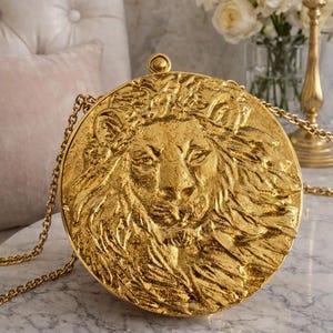 May include: A round, gold-colored clutch bag featuring a highly detailed, three-dimensional embossed lion head on the front surface. The bag includes a thin, matching gold-tone metal chain strap and a small, circular clasp at the top.