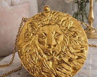 May include: A round, gold-colored clutch bag featuring a highly detailed, three-dimensional embossed lion head on the front surface. The bag includes a thin, matching gold-tone metal chain strap and a small, circular clasp at the top.