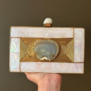 Puede incluir: Un bolso de mano rectangular con incrustaciones de concha blanca iridiscente enmarcado por detalles de alambre de metal dorado. Una piedra de ágata gris pulida se sitúa en el centro y un cierre de piedra blanca corona el borde superior.