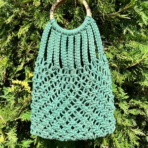 May include: A green macrame bag with a wooden handle. The bag is made of knotted rope and has a unique, intricate design.