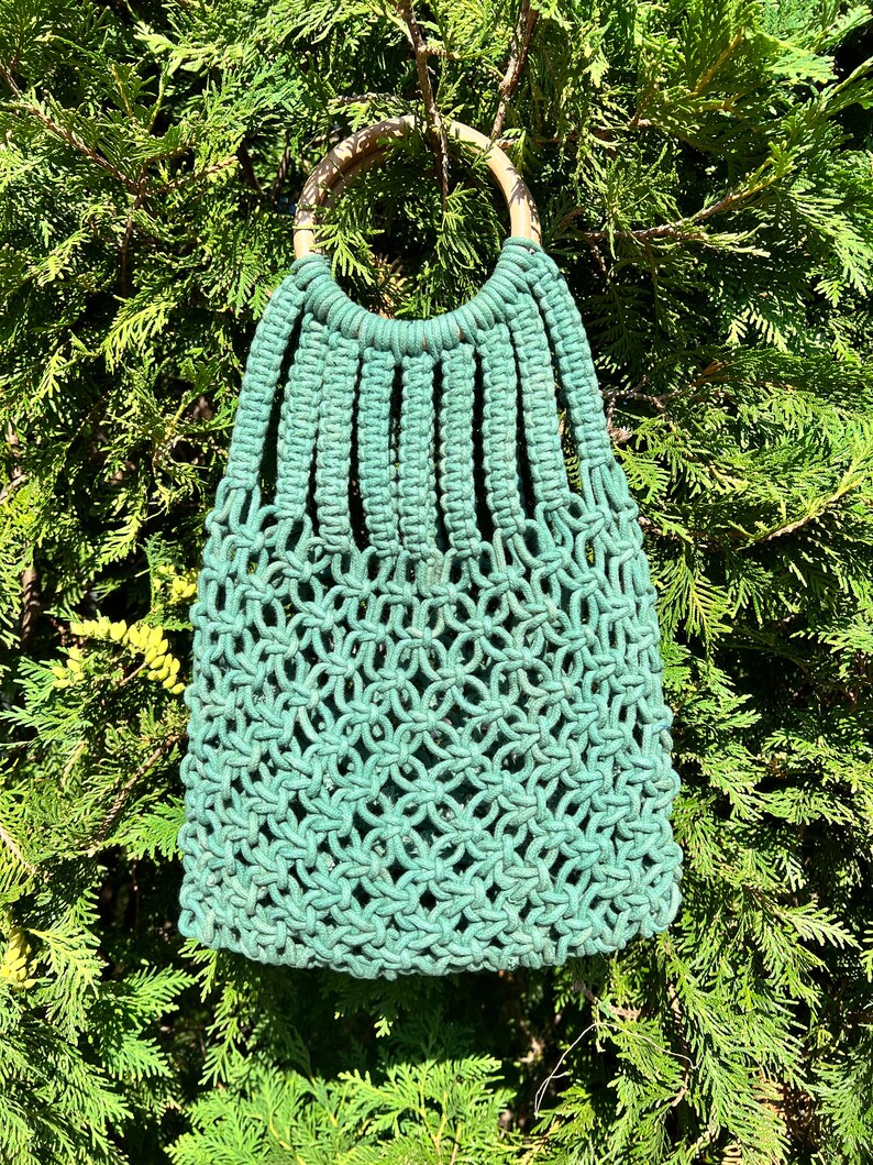 May include: A green macrame bag with a wooden handle. The bag is made of knotted rope and has a unique, intricate design.