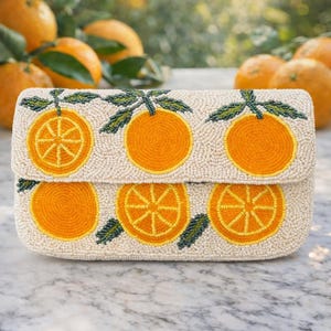 May include: Cream-colored beaded clutch featuring a repeating pattern of orange slices and whole oranges with green leaves. The clutch has a rectangular shape with a flap closure. The background shows oranges on a tree.