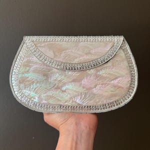 Puede incluir: Un bolso de mano nacarado con ribete plateado. El bolso tiene una solapa curva y un borde plateado decorativo. La superficie iridiscente refleja tonos rosados, verdes y blancos. El bolso se sostiene sobre un fondo oscuro.