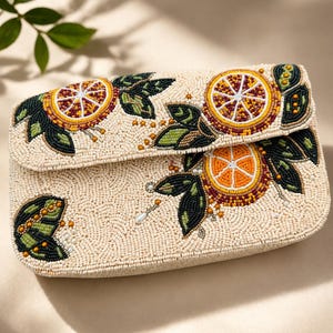 May include: A rectangular clutch bag covered in cream-coloured beads featuring a detailed pattern of orange slices and dark green leaves. The citrus design is rendered in vibrant orange, yellow, and deep red beads, accented with small white pearl-like beads.