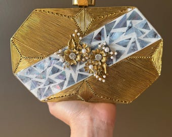 May include: A gold and white clutch bag with a geometric design and a floral embellishment. The clutch has a hinged closure and a decorative top.