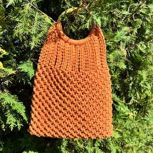 May include: A brown crocheted tote bag with a wooden handle. The bag is hanging from a tree branch.