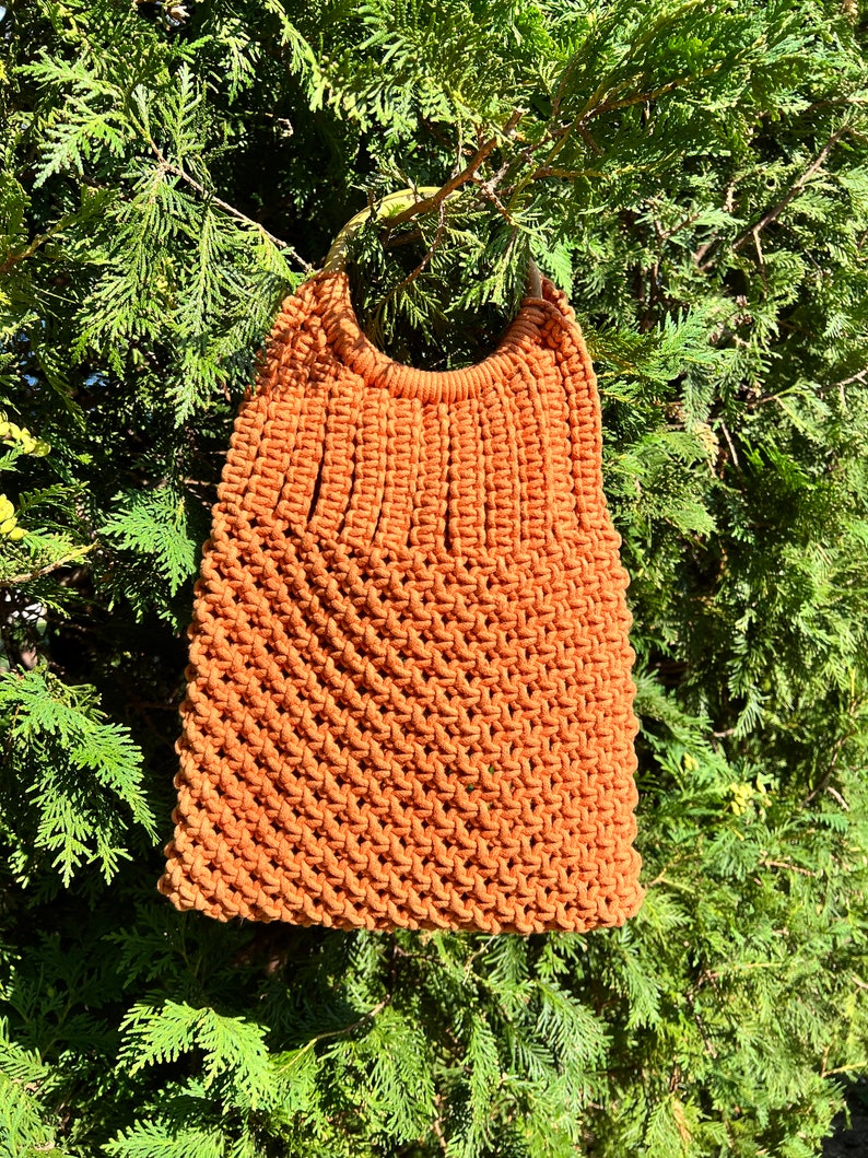 May include: A brown crocheted tote bag with a wooden handle. The bag is hanging from a tree branch.