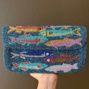 Puede incluir: Un bolso de mano rectangular cubierto de cuentas azul oscuro con un patrón de cuentas multicolor de varios peces. Los peces están representados en tonos naranja, amarillo, azul claro, rosa, azul oscuro, morado y blanco, creando una escena acuática.