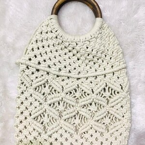 May include: A white crocheted tote bag with a wooden handle. The bag has a geometric pattern and is made of thick rope.