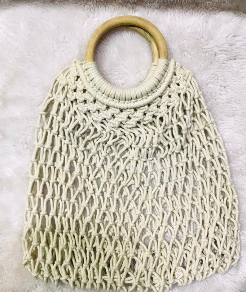 May include: A white crocheted tote bag with a wooden handle. The bag is made of a thick, rope-like material and has a wide, open top.