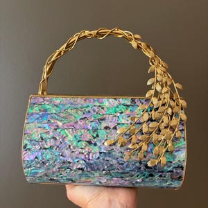 May include: A rectangular clutch bag featuring a mosaic of iridescent abalone shell tiles in shades of blue, green, and purple. The bag has a gold-toned metal frame and a curved handle adorned with a decorative gold-toned metal leaf branch.