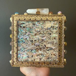 以下が含まれることがあります： 正方形の金色の箱で、装飾的なフレームと虹色のあわびの貝殻の象嵌が施されています。箱はカラフルな宝石と透明なクリスタルで飾られています。箱は手で持たれています。