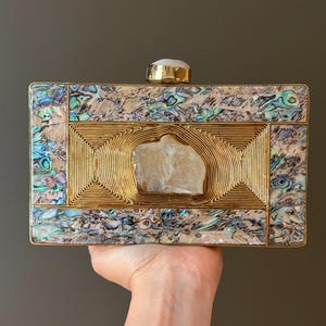 May include: A rectangular clutch bag featuring an iridescent abalone shell inlay border and a central gold-toned metal panel with concentric line patterns. A rough-cut, translucent white stone is embedded in the center, with a matching stone clasp.