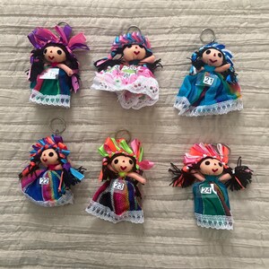May include: Six doll keychains with black yarn hair, colorful ribbon headpieces, and fabric dresses featuring lace trim. Each doll has a small white tag numbered 19, 20, 21, 22, 23, or 24 attached to the front of the dress, and a metal ring.