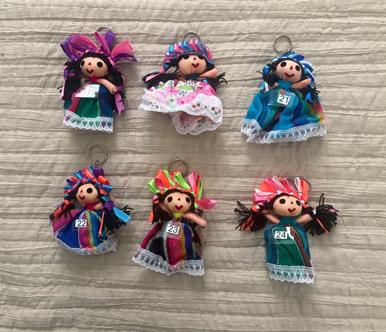 May include: Six doll keychains with black yarn hair, colorful ribbon headpieces, and fabric dresses featuring lace trim. Each doll has a small white tag numbered 19, 20, 21, 22, 23, or 24 attached to the front of the dress, and a metal ring.