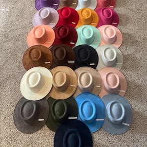 May include: A collection of 25 wide-brimmed hats in various colors, including brown, beige, purple, red, green, blue, and black. The hats are made of a soft, suede-like material and have a flat top.